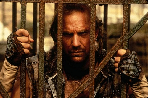 Kevin Costner in the movie Water world looking through the metal bars of the cage he is locked in.